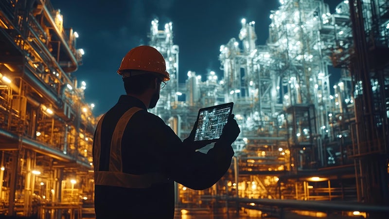 A worker in a hard hat uses a tablet in an illuminated industrial setting at night