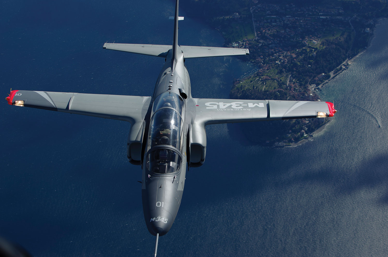 A military jet aircraft is shown in flight over a large body of water, viewed from above