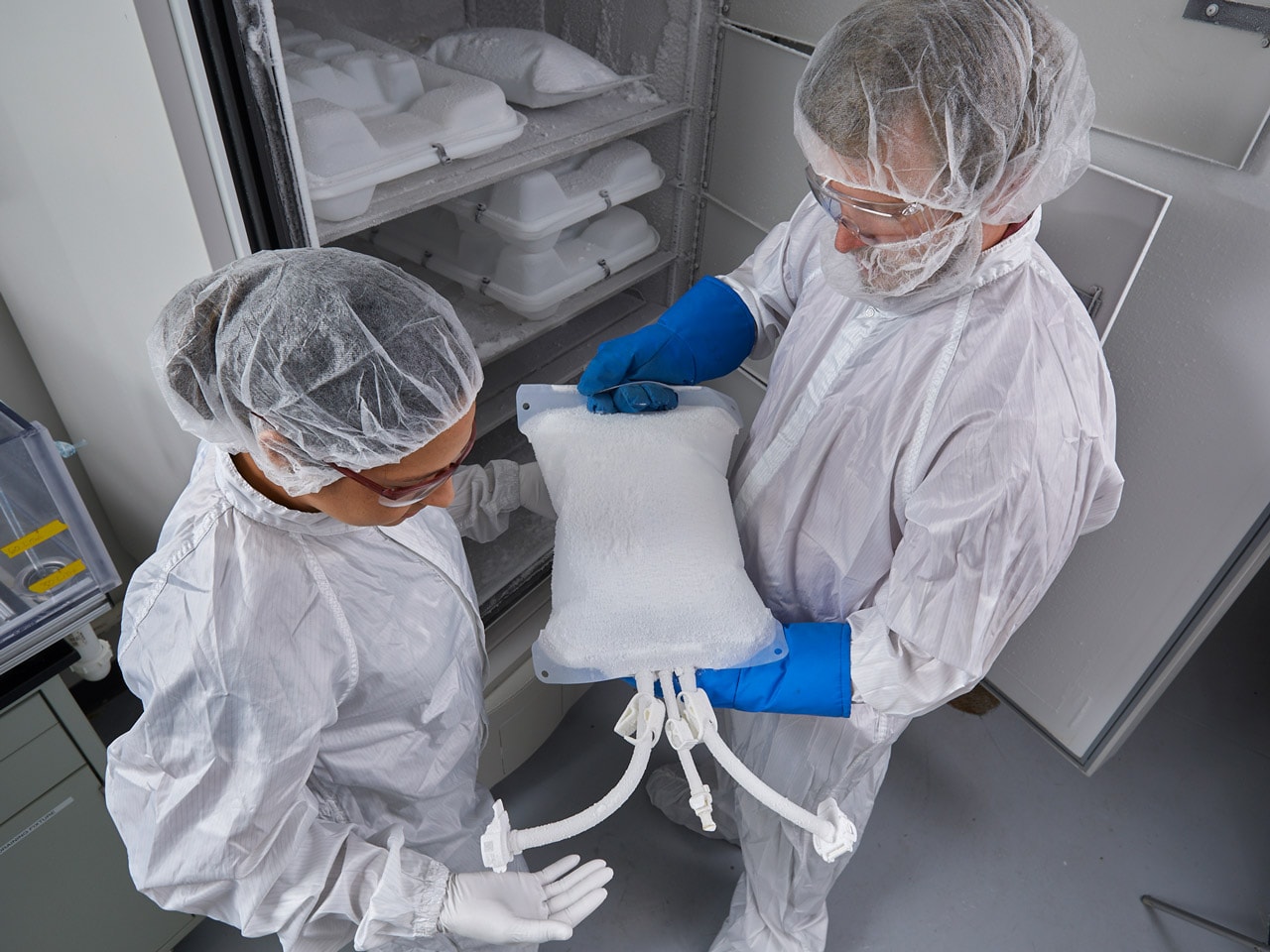 Two laboratory scientists wearing lab coats, safety goggles, and gloves adjust tubing and a sample vial on a Bio-Rad chromatography system, focusing on the instrument’s column and control modules in a research lab setting.