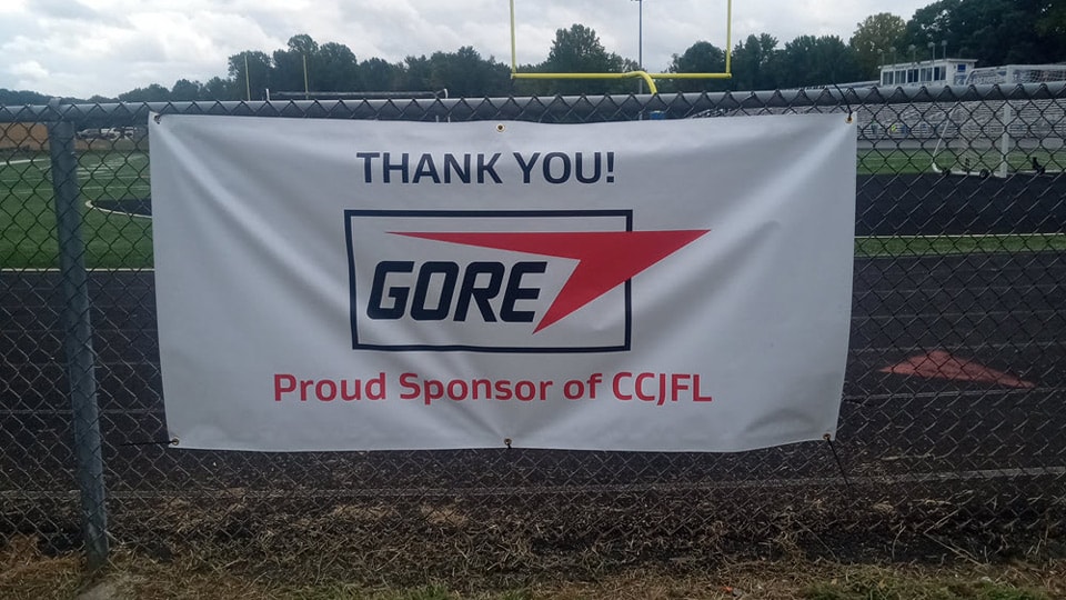 Banner attached to a chain-link fence at a football field reads, “THANK YOU! GORE Proud Sponsor of CCJFL.”