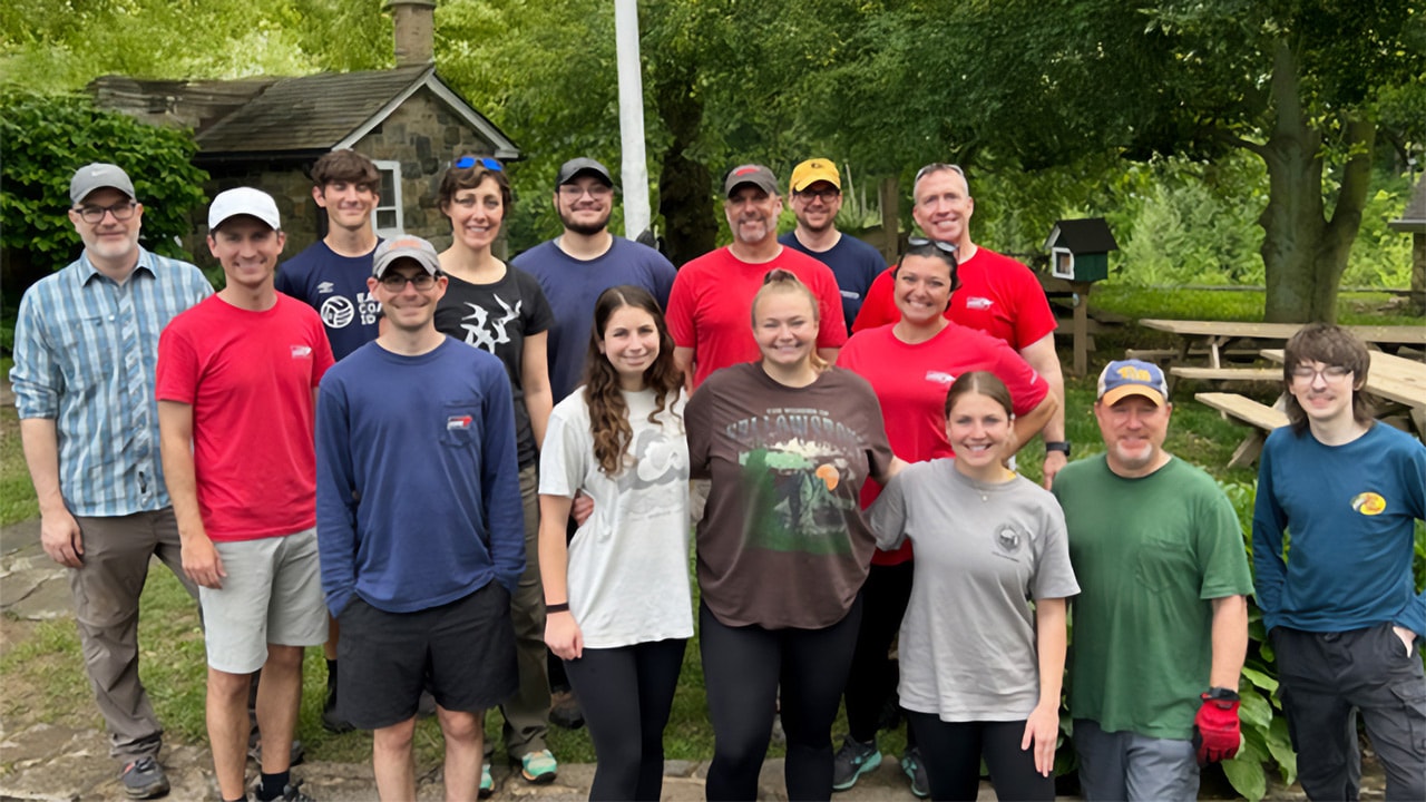 A group of volunteers pose together outdoors in a park-like setting with trees, picnic tables, and a small stone building in the background.