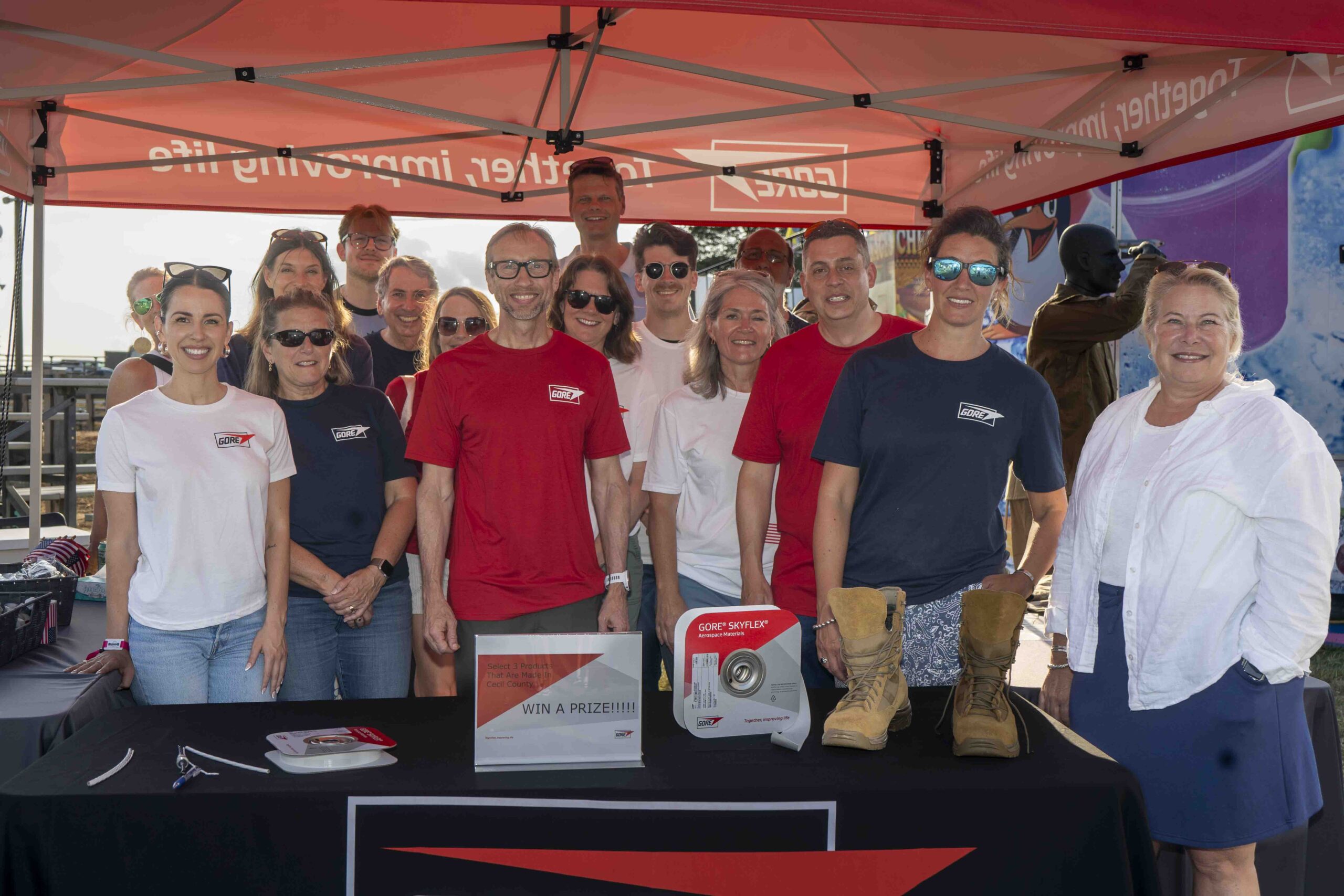A group of people wearing GORE shirts pose together under a red GORE canopy at an outdoor event booth, with promotional materials and a pair of boots displayed on the table in front of them.