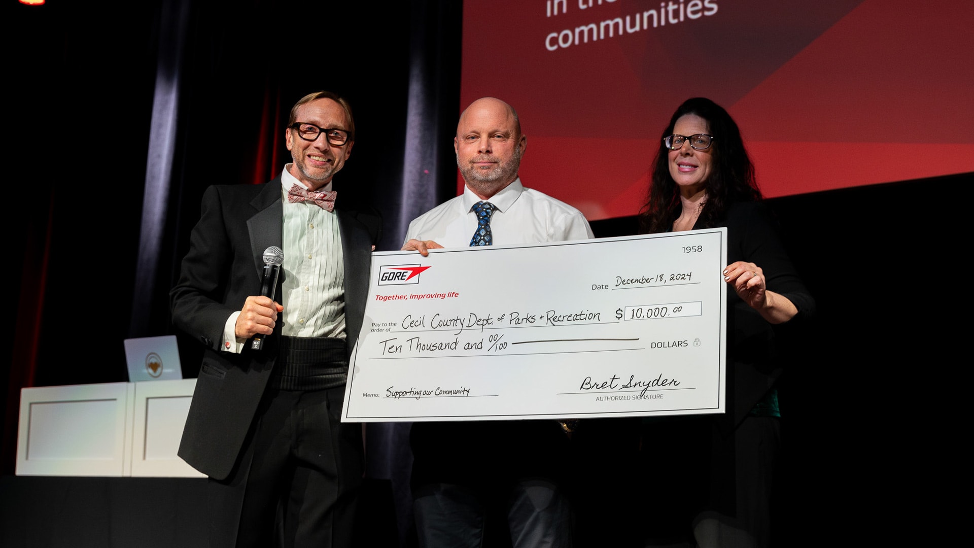 Three people stand on a stage holding an oversized $10,000 donation check from GORE made out to the Cecil County Department of Parks and Recreation, dated December 18, 2024.