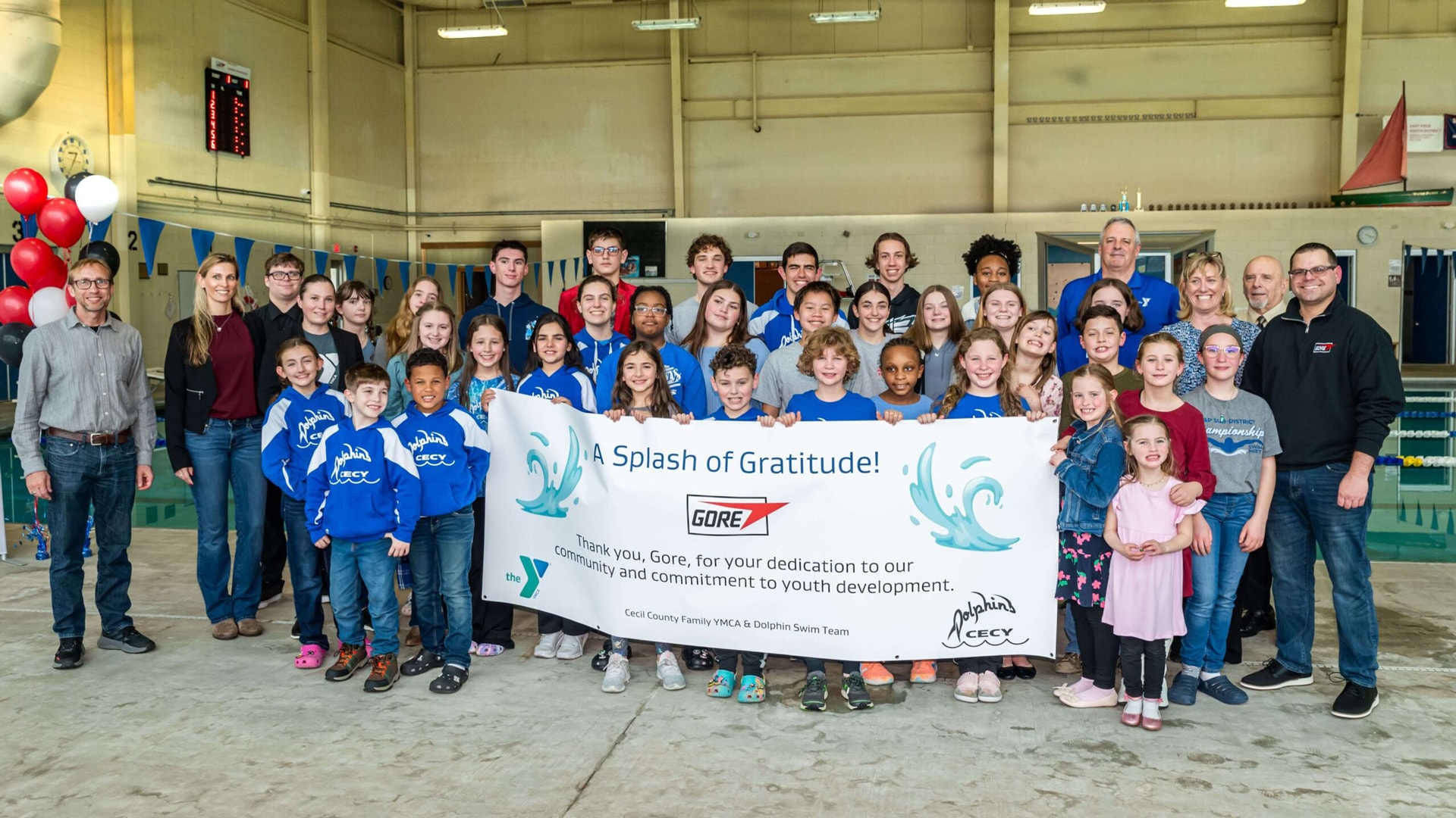 A large group of children and adults stand together inside an indoor pool facility holding a banner that reads, “A Splash of Gratitude! Thank you, Gore, for your dedication to our community and commitment to youth development,” from the Cecil County Family YMCA and Dolphin Swim Team.