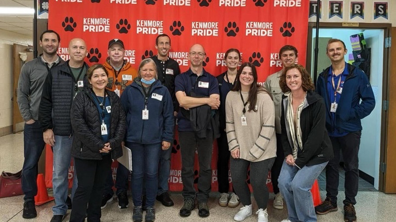A group of employees pose in front of a red “Kenmore Pride” backdrop decorated with paw print graphics inside a school hallway.