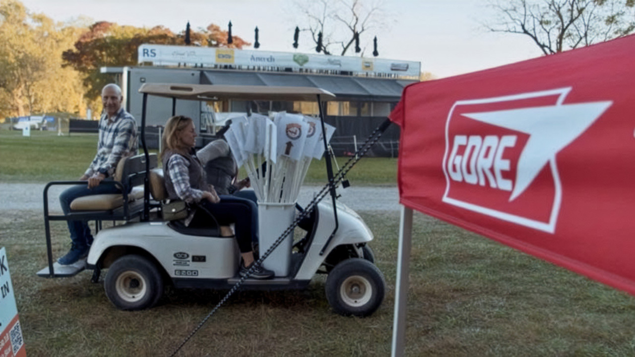 Two people ride in a golf cart carrying white course signs at an outdoor event, with a red GORE banner in the foreground and a stage structure in the background.