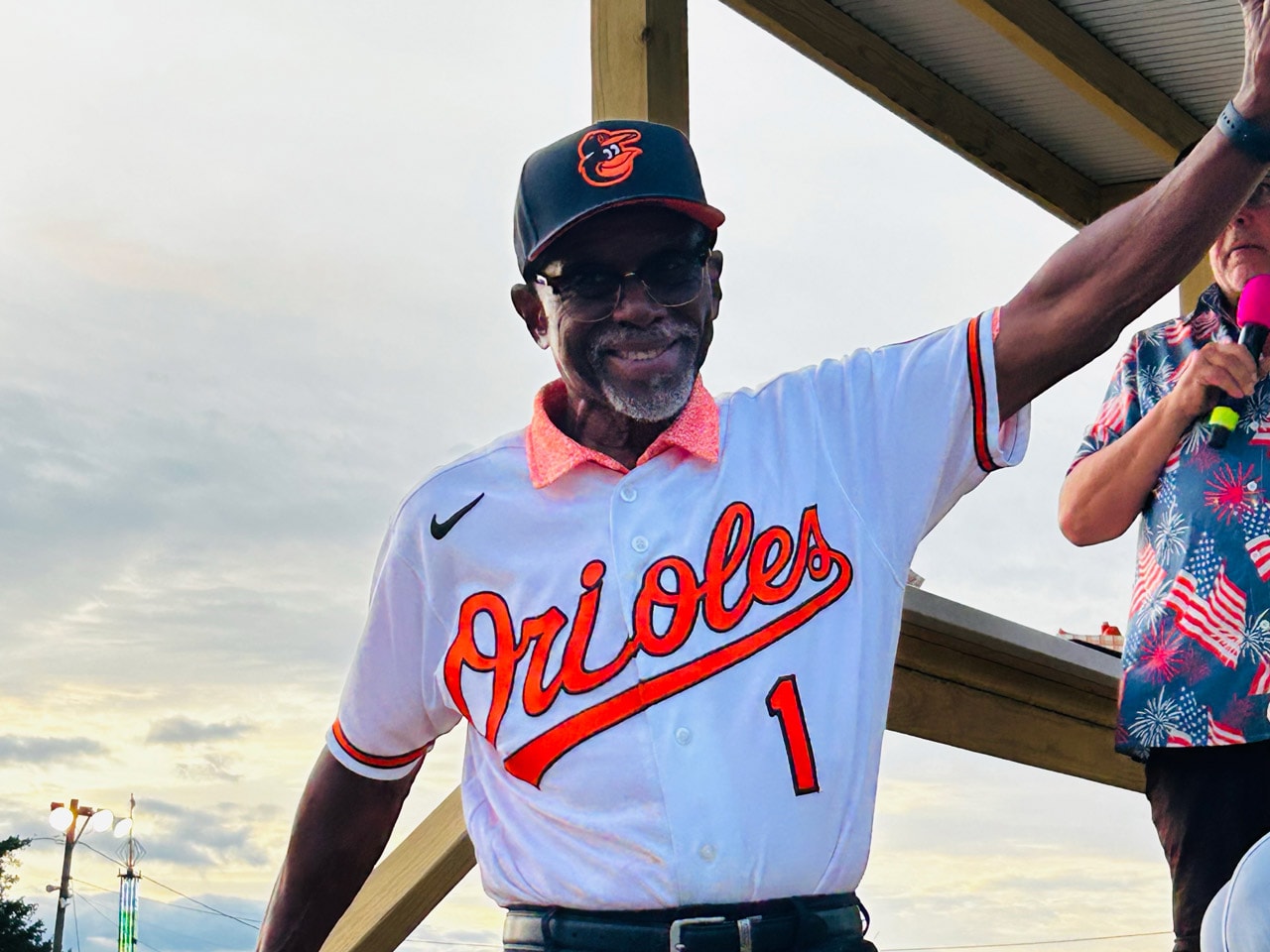 Al Bumbry, wearing a Baltimore Orioles jersey, waves to the crowd from a stage during Military Appreciation Day while an announcer stands nearby with a microphone.