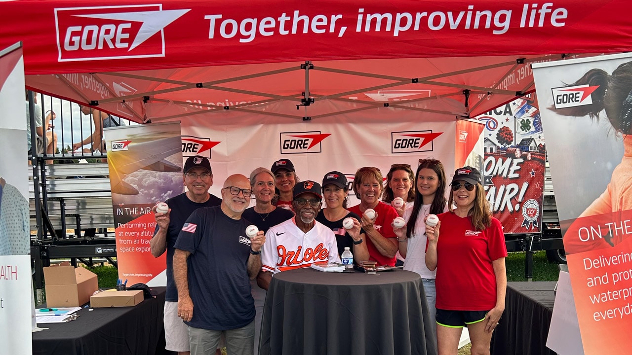 Group photo of Hall of Fame Baltimore Orioles player Al Bumbry and Gore team members under a red Gore event tent at the Cecil County Fair, smiling and holding autographed baseballs. Gore banners and the slogan “Together, improving life” are displayed throughout the booth.