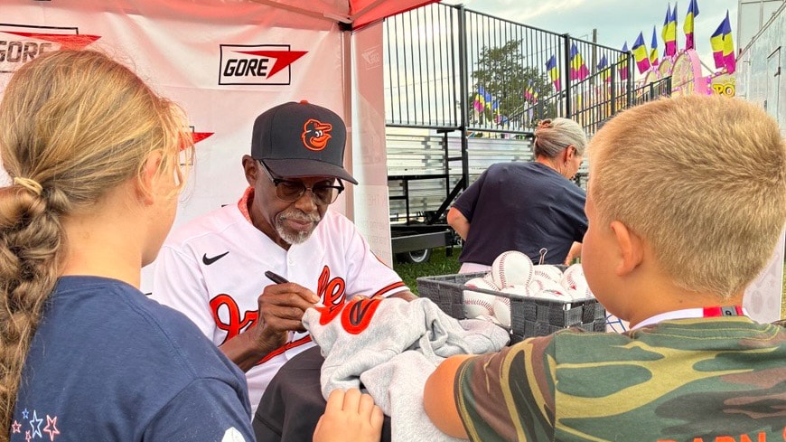 Al Bumbry signs memorabilia for children at the Gore booth during the Cecil County Fair, seated at a table with baskets of baseballs ready for autographs.