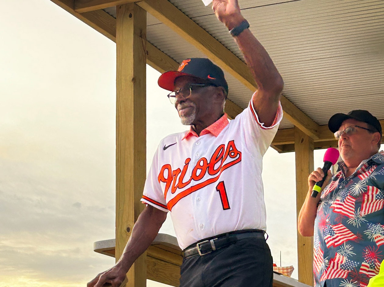 Al Bumbry waves to the audience from a stage during Military Appreciation Day as an announcer with a microphone stands beside him against an evening sky.