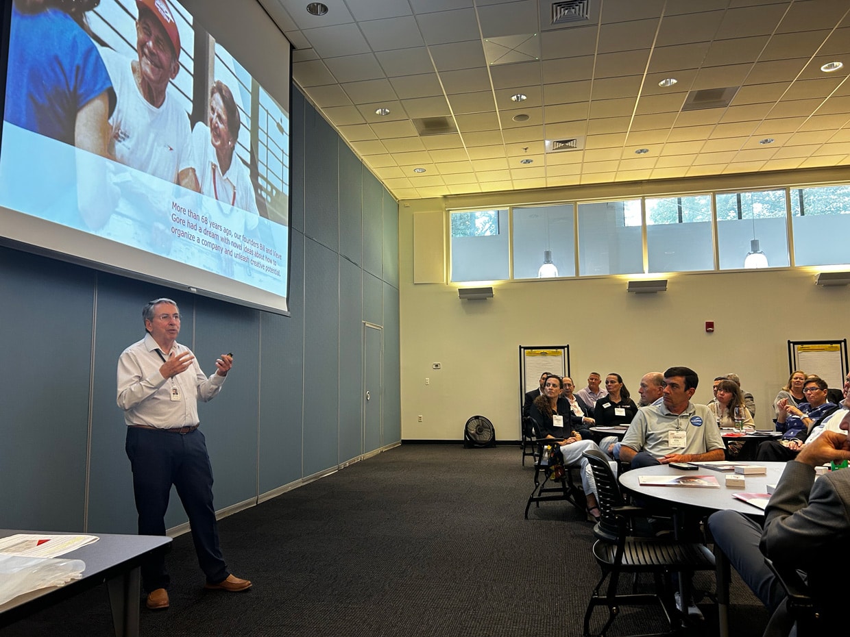 A male presenter speaks to attendees seated at round tables during a W. L. Gore & Associates event, with a large screen displaying a presentation about the company’s history and founding vision.