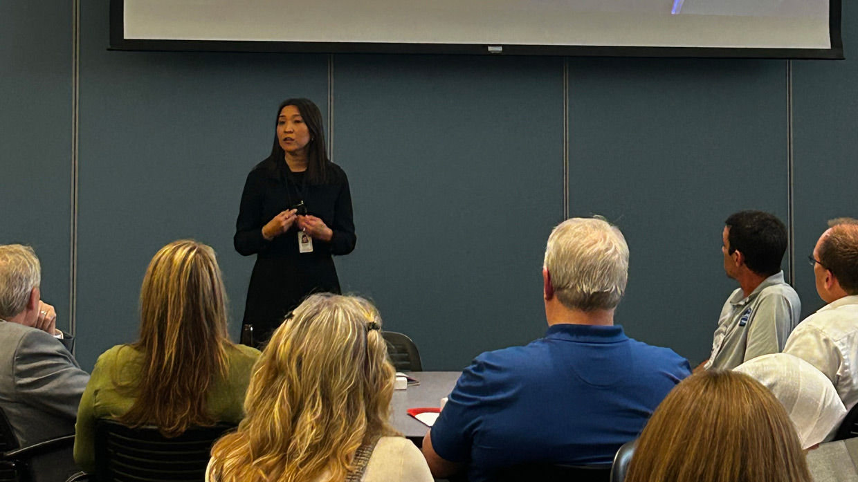 A woman delivers a presentation to a seated audience in a conference room, speaking and gesturing as attendees listen during a group meeting.