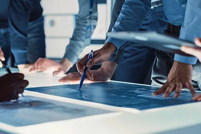 A busy meeting with several people in business attire; the focus is a man's hands drawing a diagram