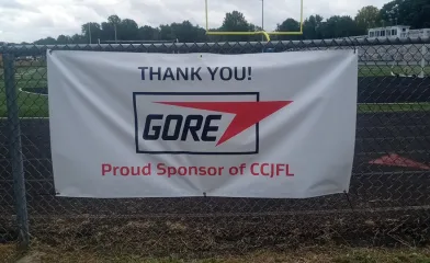 Banner attached to a chain-link fence at a football field reads, “THANK YOU! GORE Proud Sponsor of CCJFL.”