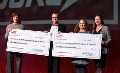 Four people stand on a stage holding oversized donation checks at a GORE event. One check is for $10,000 to Friends of the Newark Free Library, and the other is for $10,000 to Friends of Cecil County Public Library, both dated 12/5/2025 for “2025 Holiday Party Donation.”