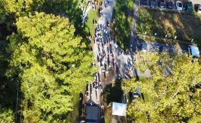 Aerial view of a large group of people walking along a paved path through a wooded park area, with parked cars and event tents visible nearby.