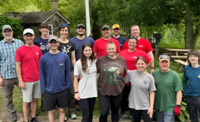 A group of volunteers pose together outdoors in a park-like setting with trees, picnic tables, and a small stone building in the background.