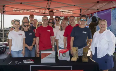 A group of people wearing GORE shirts pose together under a red GORE canopy at an outdoor event booth, with promotional materials and a pair of boots displayed on the table in front of them.