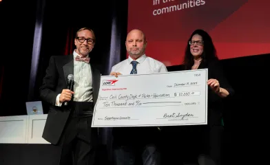 Three people stand on a stage holding an oversized $10,000 donation check from GORE made out to the Cecil County Department of Parks and Recreation, dated December 18, 2024.