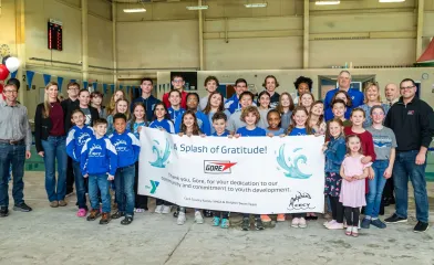A large group of children and adults stand together inside an indoor pool facility holding a banner that reads, “A Splash of Gratitude! Thank you, Gore, for your dedication to our community and commitment to youth development,” from the Cecil County Family YMCA and Dolphin Swim Team.