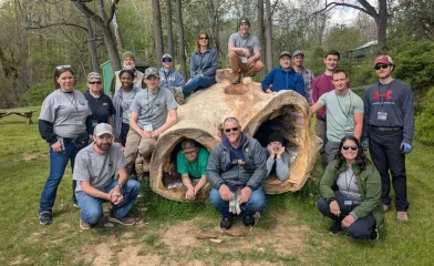 Group of volunteers posing outdoors around a large hollow tree trunk in a wooded park area during a community cleanup or Earth Day event.