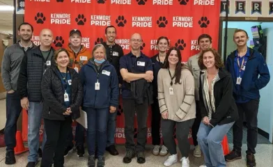 A group of employees pose in front of a red “Kenmore Pride” backdrop decorated with paw print graphics inside a school hallway.