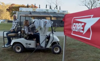 Two people ride in a golf cart carrying white course signs at an outdoor event, with a red GORE banner in the foreground and a stage structure in the background.