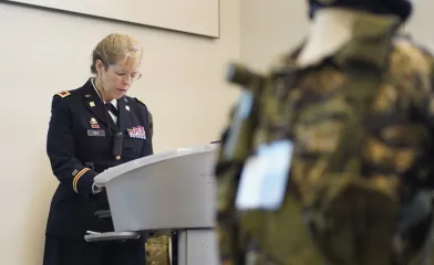 A woman in a decorated military dress uniform stands at a podium reading from prepared remarks, while a blurred camouflage uniform is visible in the foreground.