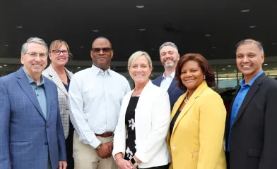 Seven professionally dressed adults stand together outside a building entrance, smiling for a group photo.