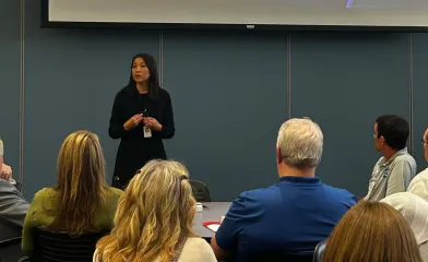 A woman delivers a presentation to a seated audience in a conference room, speaking and gesturing as attendees listen during a group meeting.