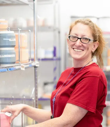 Smiling woman in a red shirt and glasses organizing containers on metal shelving in a brightly lit storage or manufacturing facility.