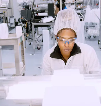 An Associate in cleanroom gear inspects material on a roll in a high-tech lab or manufacturing facility, surrounded by equipment and packaging supplies.